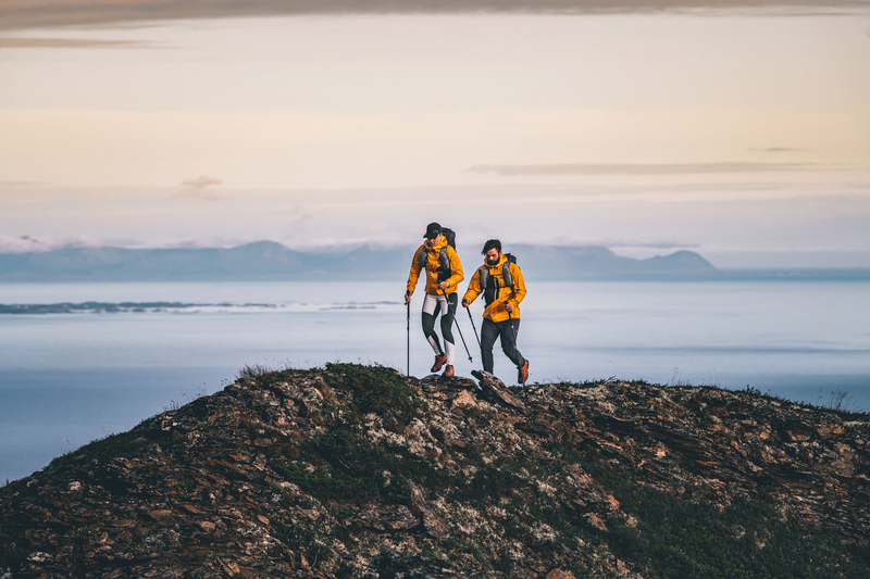 Två personer vandrar på ett berg i bakgrunden syns hav och berg